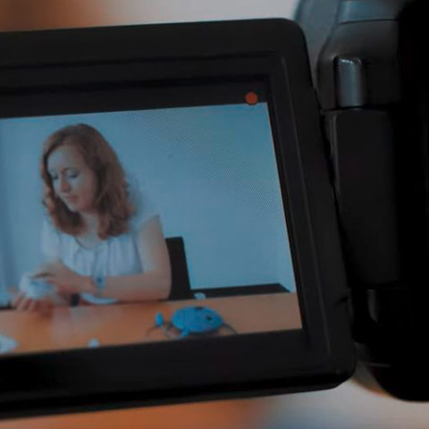 A video camera screen shows a young woman sitting at a table, working with small white objects.