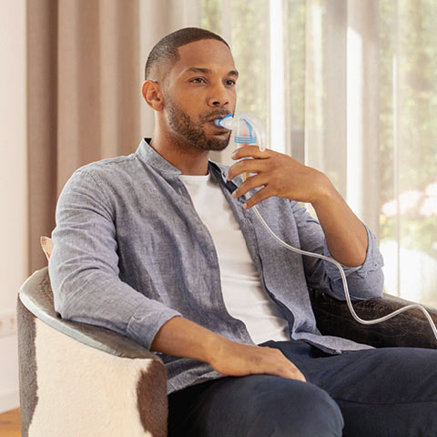 A young man inhales relaxed in a living room armchair using a PARI inhalation device