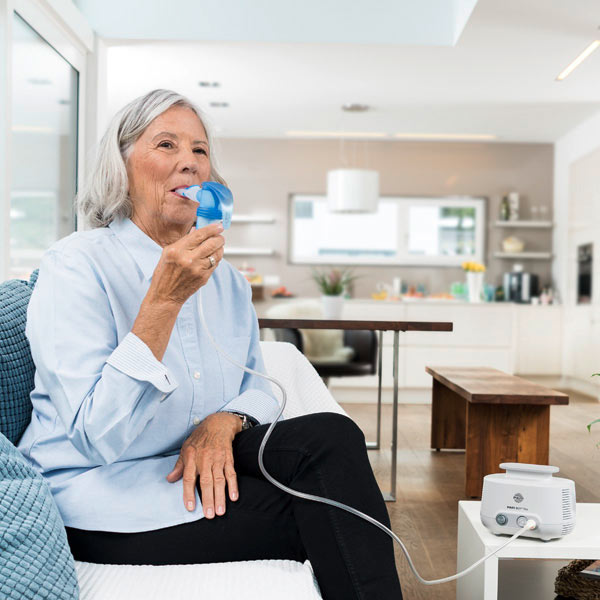 An elderly woman sits relaxed on a sofa in a living room. She uses a PARI LC SPRINT nebulizer connected via a tube to the PARI COMPACT2 compressor on the table.