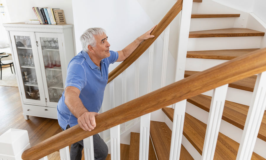 An elderly man slowly climbs a wooden staircase. He holds onto the railing with both hands and appears exhausted or short of breath.