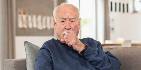 An elderly man sits on a sofa in a living room. He wears a dark blue sweater and has a thoughtful expression as he coughs into his hand.