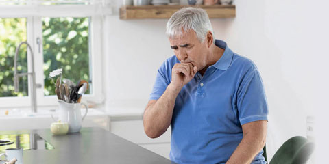 An older man with gray hair and a blue polo shirt stands in a bright kitchen and coughs into his hand.