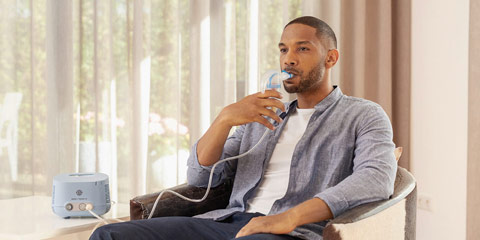 A man sits relaxed in an armchair and uses a PARI COMPACT2 inhaler. He holds a mouthpiece connected to a tube to his mouth, while the blue inhaler sits on a table next to him.
