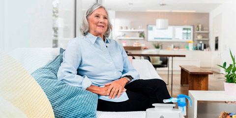 Older woman with grey hair sits relaxed on a white sofa and looks relaxed into the room.