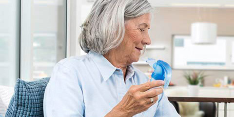 An elderly woman sits on a sofa in a modern, bright room. She holds a PARI LC SPRINT nebulizer in her right hand while turning on the PARI BOY Classic with her left hand.